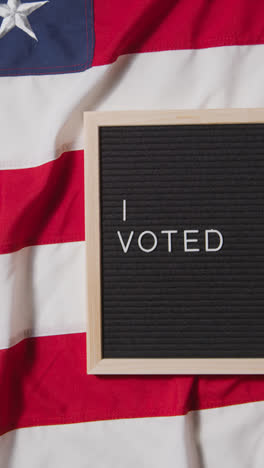 Vertical-Video-Of-Sign-Reading-I-Voted-Lying-On-US-Stars-And-Stripes-Flag-For-American-Election-2024-1